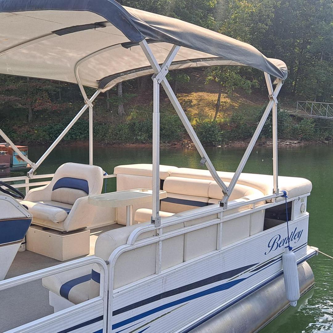 Pontoon boat with canopy docked on a calm lake, surrounded by trees.