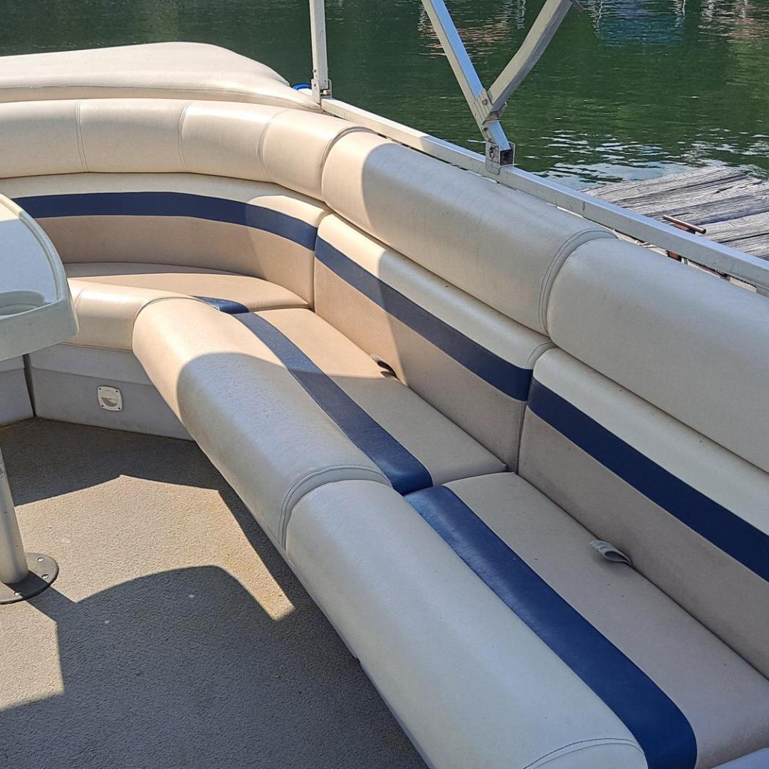 Pontoon boat interior with U-shaped beige seats and a small table, dock and water visible.