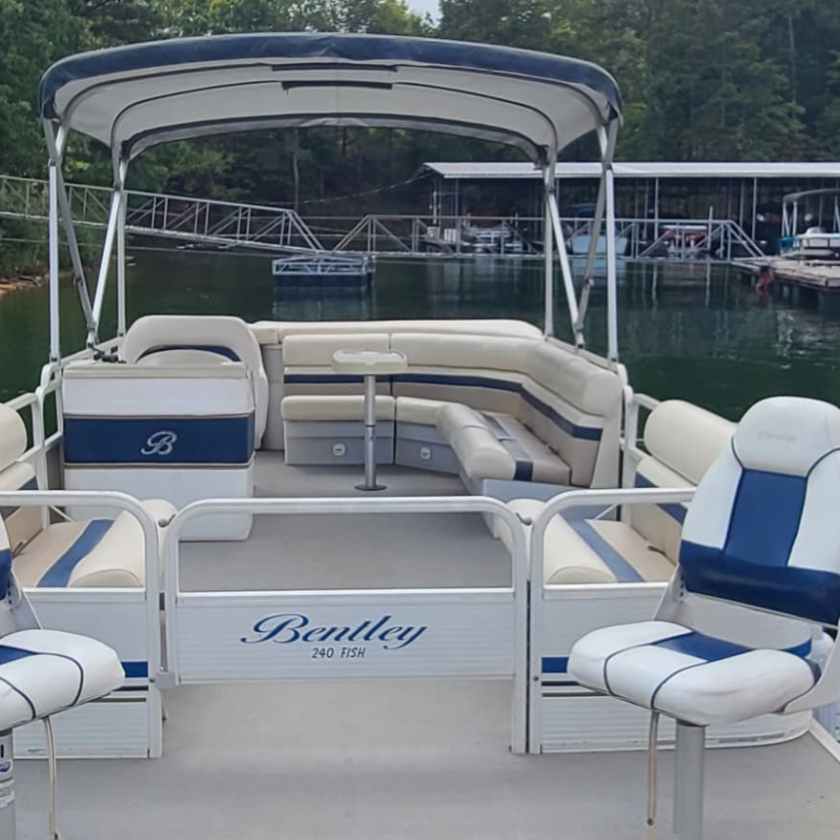 Empty pontoon boat docked near a marina with other boats and trees in the background.